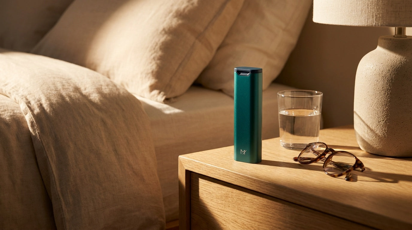A green Mannox pill box on a bedside table beside a glass of water and a pair of glasses