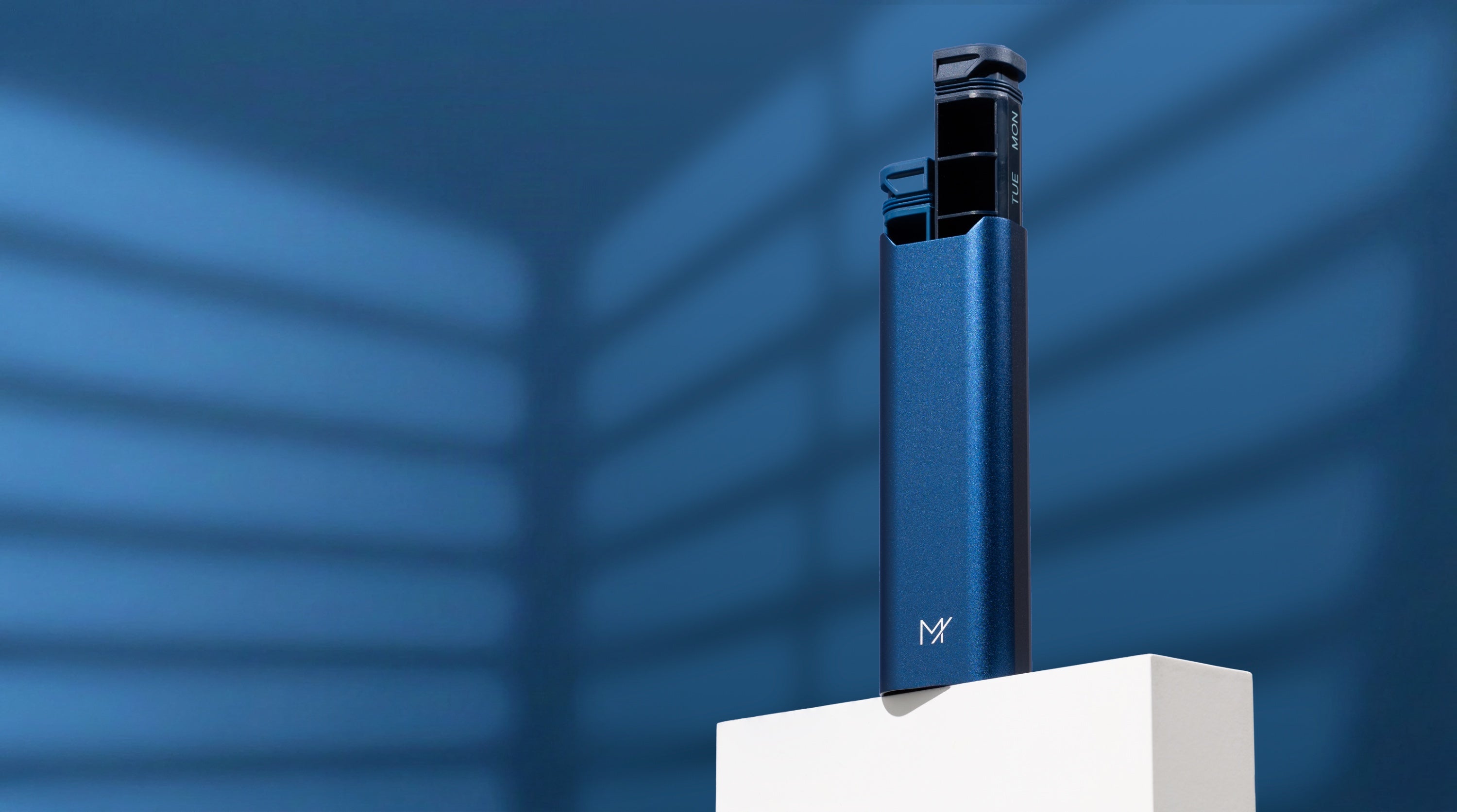 A navy Mannox AM/PM pill organiser standing part-open on a white plinth against a blue studio wall with window light