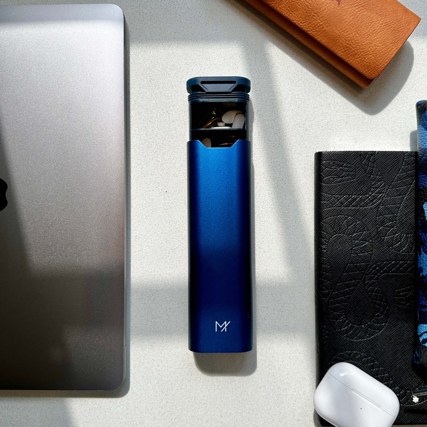 A navy Mannox Once-Daily from above, part-open with pills in the first two days, between a laptop, a passport wallet, a notebook and earphones